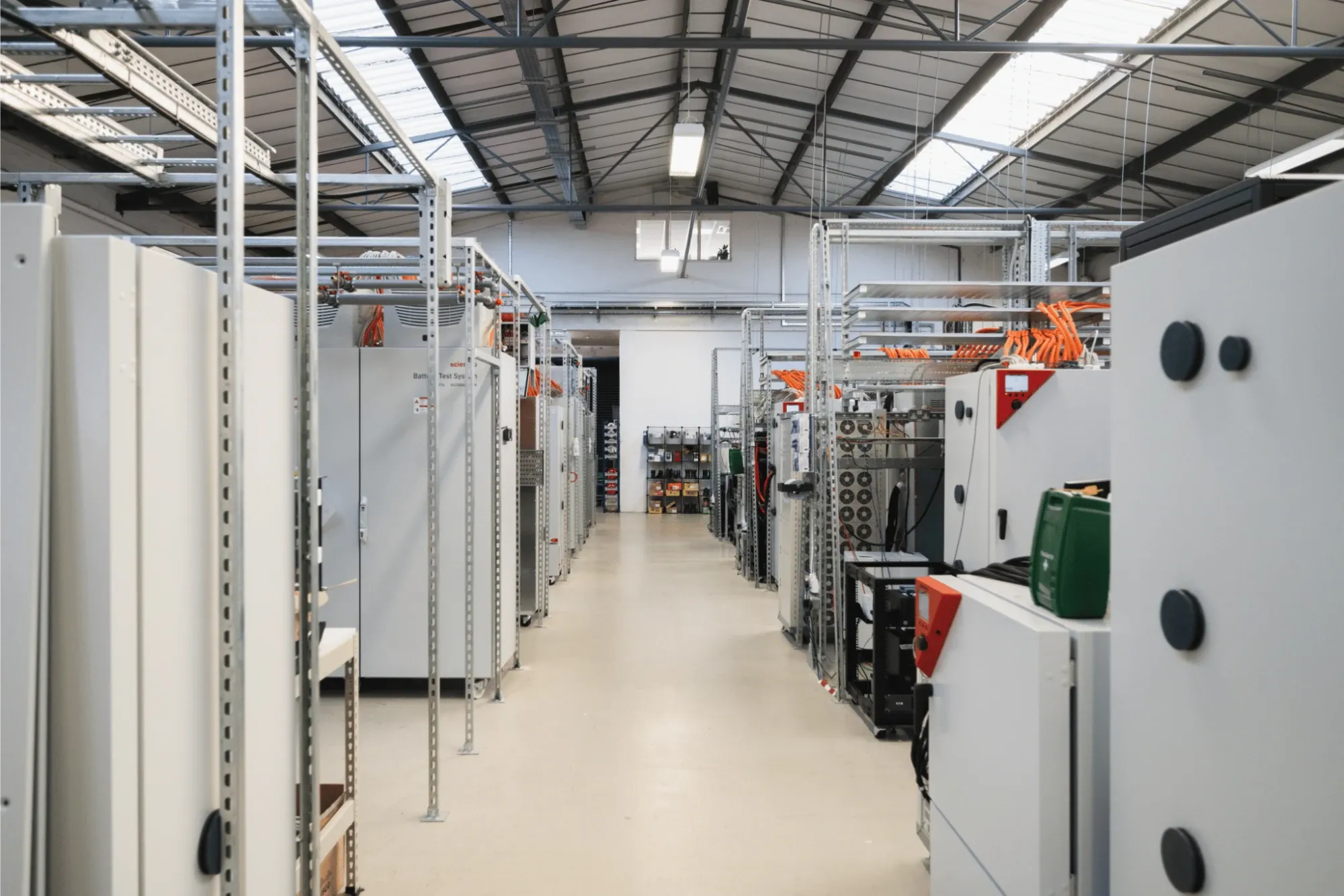 An aisle of battery cyclers and test cabinets in the battery lab, with orange cabling overhead