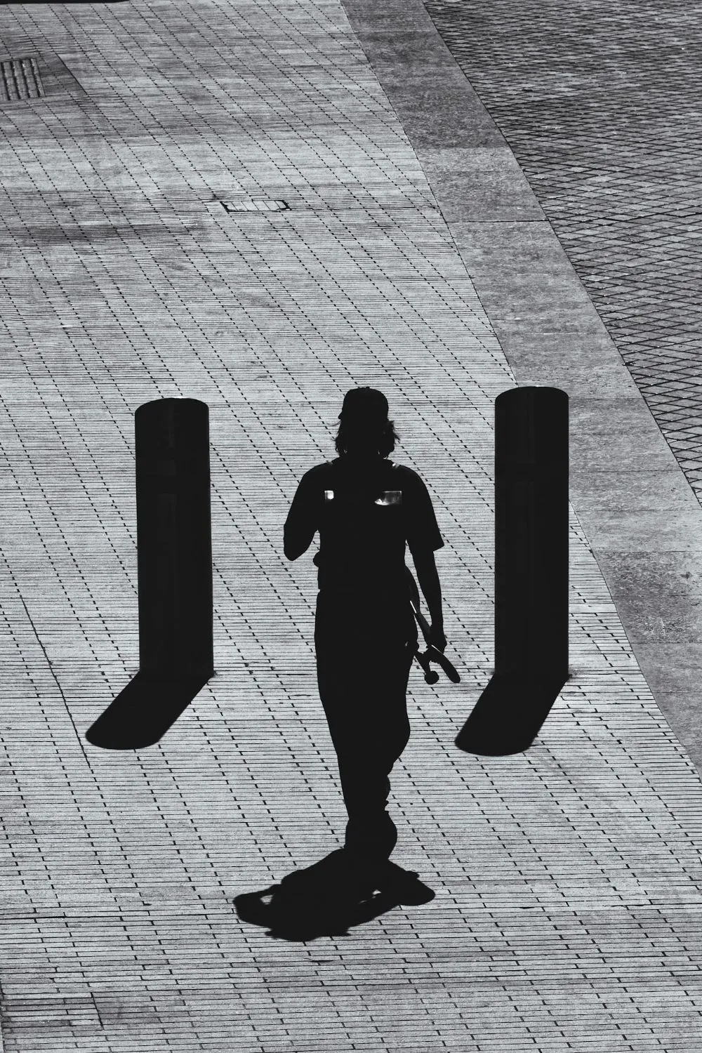 Black and white: a person in silhouette walks between two bollards on a paved square, the shadows long