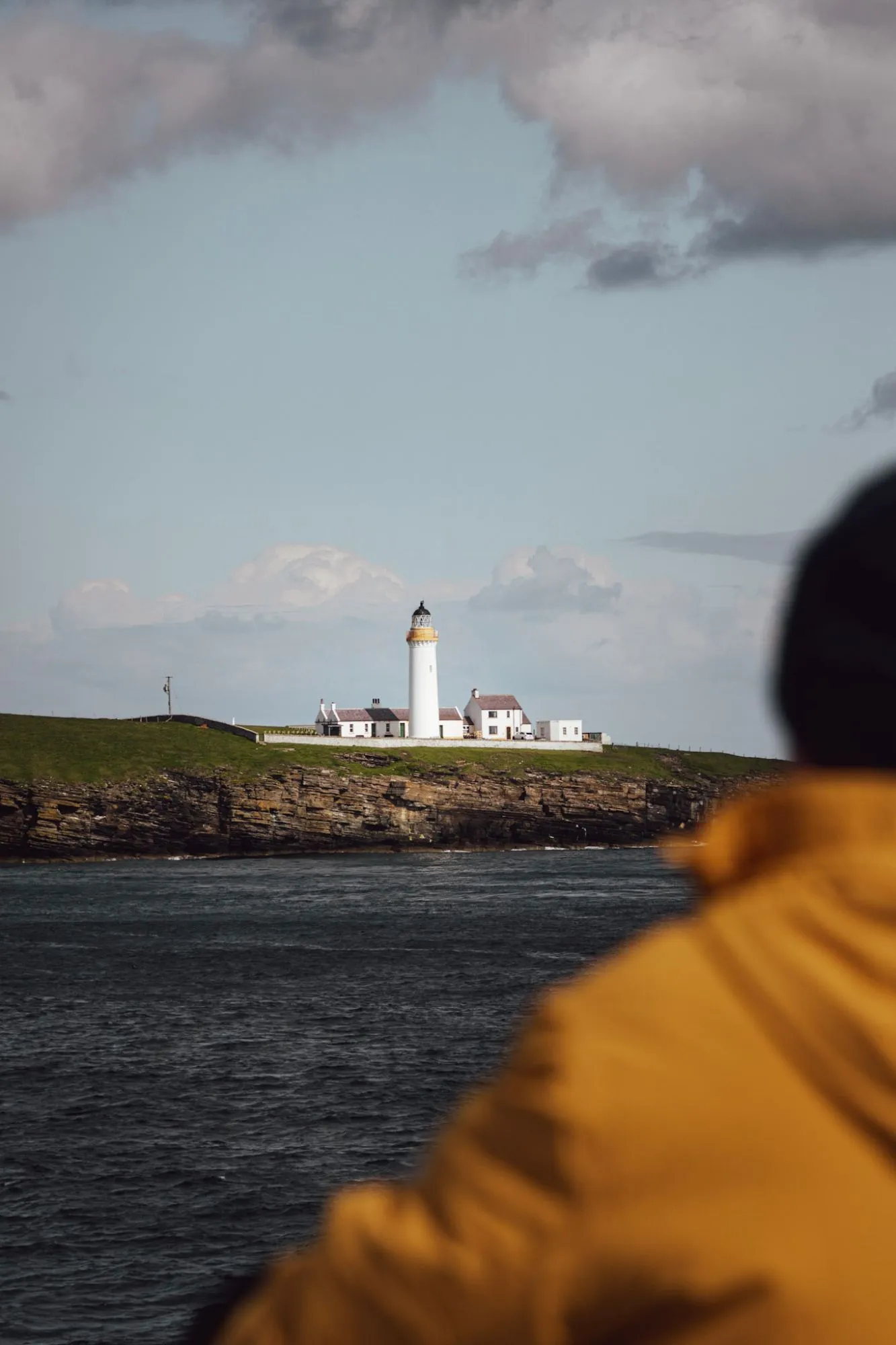 A white lighthouse and its cottages on a green headland above cliffs and dark water, a person in a yellow jacket blurred in the foreground