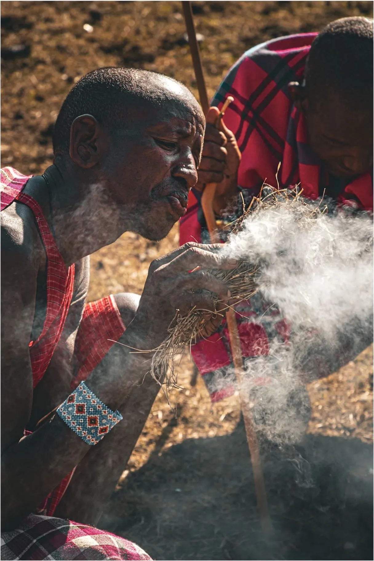 A Maasai man in red, crouching and blowing on a bundle of smoking kindling