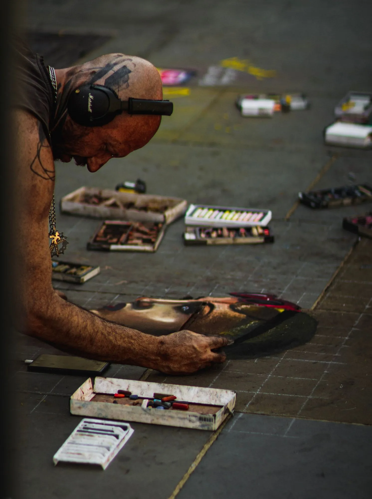 A pavement artist in headphones kneeling over a chalk portrait, boxes of pastels around him