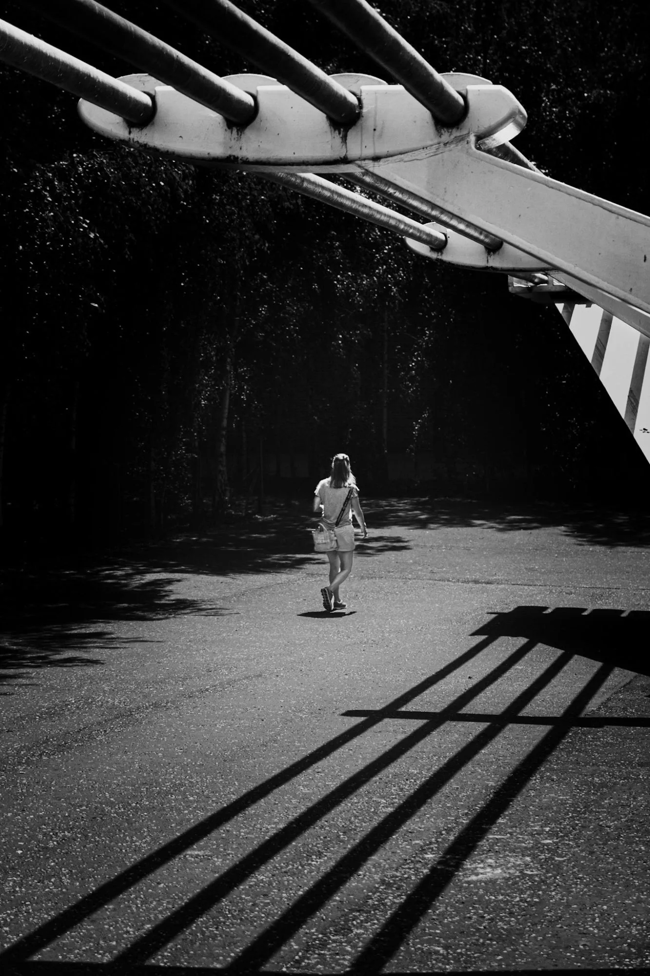 Black and white: a woman walks under the cable anchorage of a bridge, the cables striping the ground with shadow